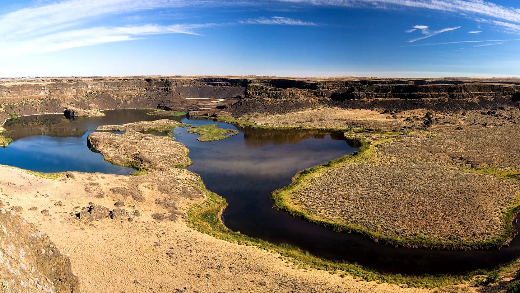 Dry Falls in Washington under a blue sky, a horseshoe of dark basalt cliffs above lakes that fill the old plunge pool, with sagebrush flats and scattered boulders to the right.