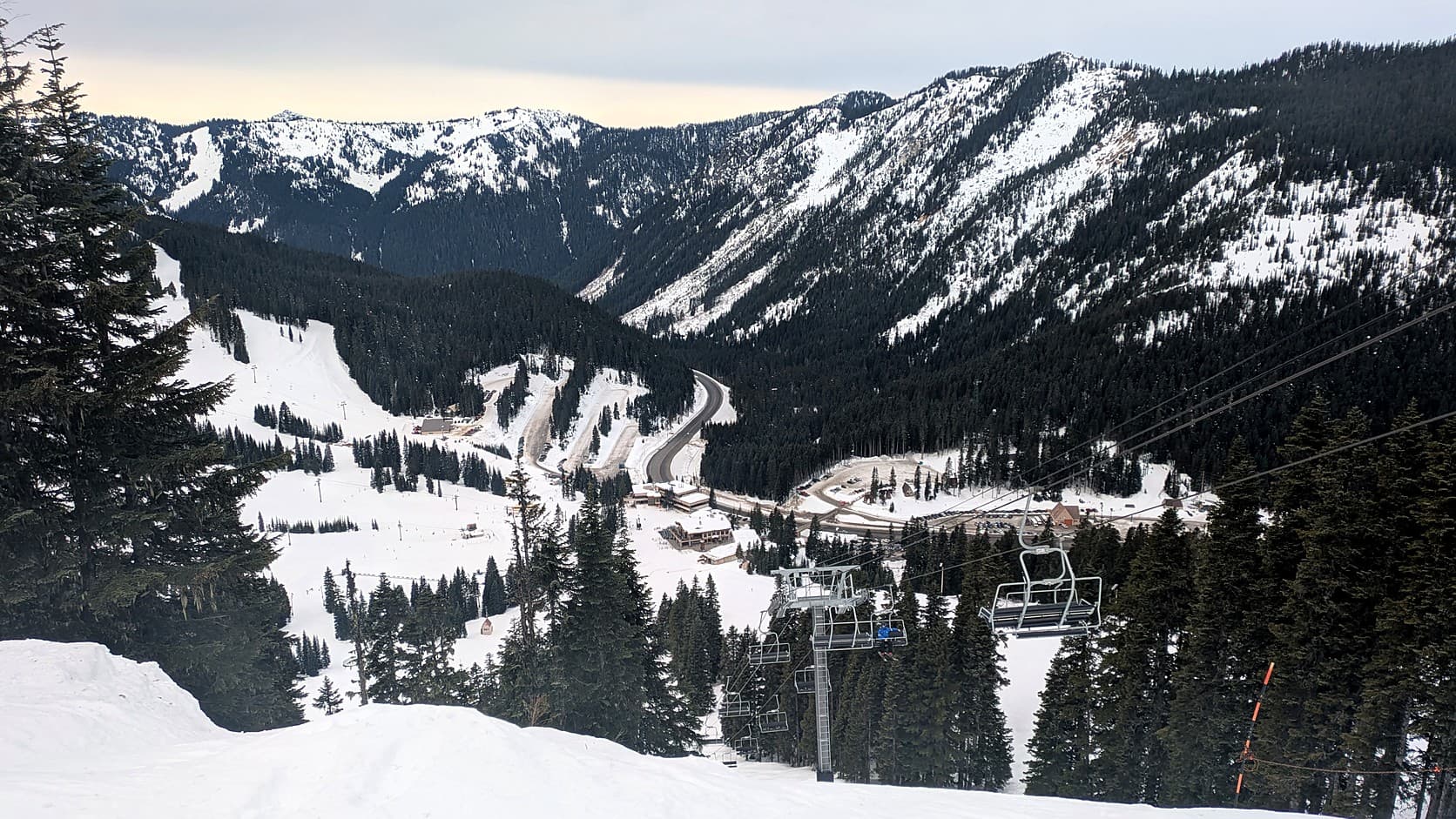 A chairlift runs down through snowy firs toward the Stevens Pass base area, with lodges, parking lots and a curving highway in the valley below and snow-streaked forested ridges under a pale sky.