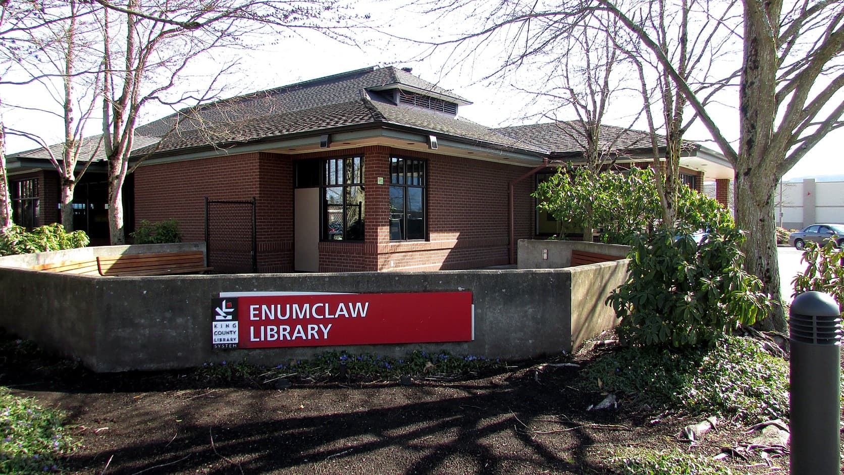 The red ENUMCLAW LIBRARY sign of the King County Library System on a low concrete wall in front of the library's brick building, with bare trees around it and a pale sky behind.