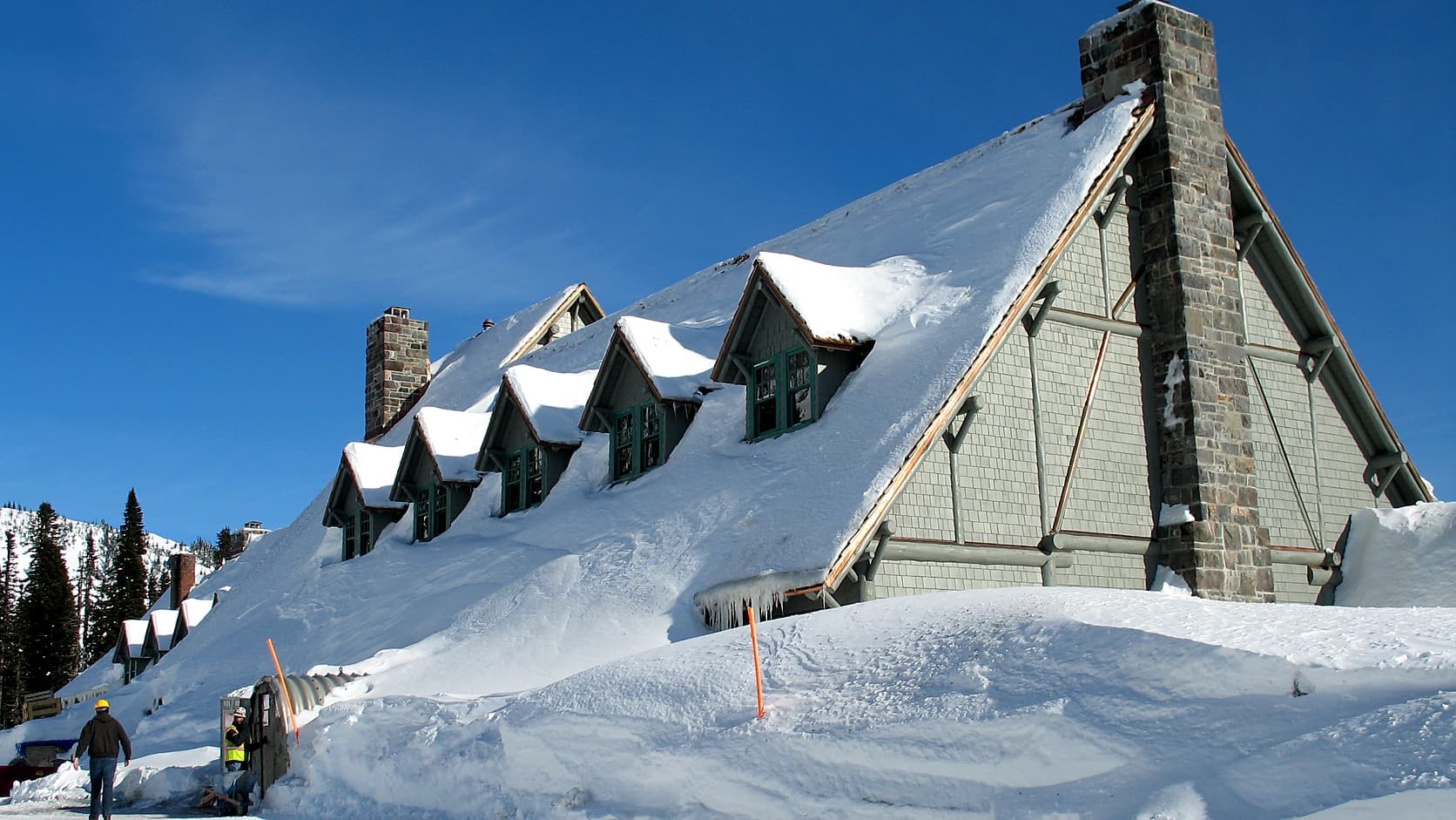 Snow drifted to the eaves of the Paradise Inn, its steep roof, row of dormer windows and stone chimney heavy with snow under a clear blue sky, with two workers in hard hats at a tunnel entrance cut into the drift.