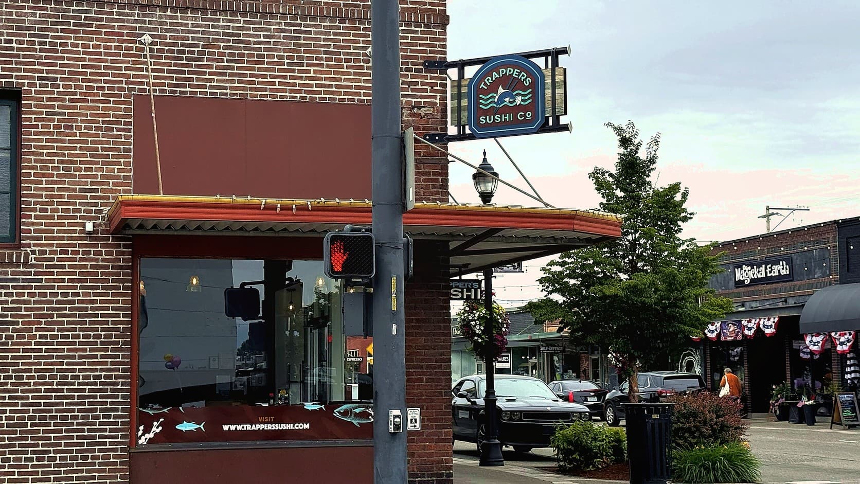 The Trapper's Sushi Co. sign, with its fish and chopsticks logo, hangs from the red-brick corner of the restaurant's building above an awning and a window lettered www.trapperssushi.com, with a crosswalk signal pole in front and parked cars and shops along Cole Street beyond.