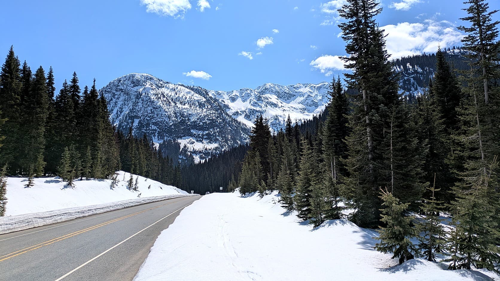 A cleared two-lane highway with a double yellow line runs between a bank of old snow and a wide snowfield toward snow-covered peaks, with dark firs on both sides under a blue sky.