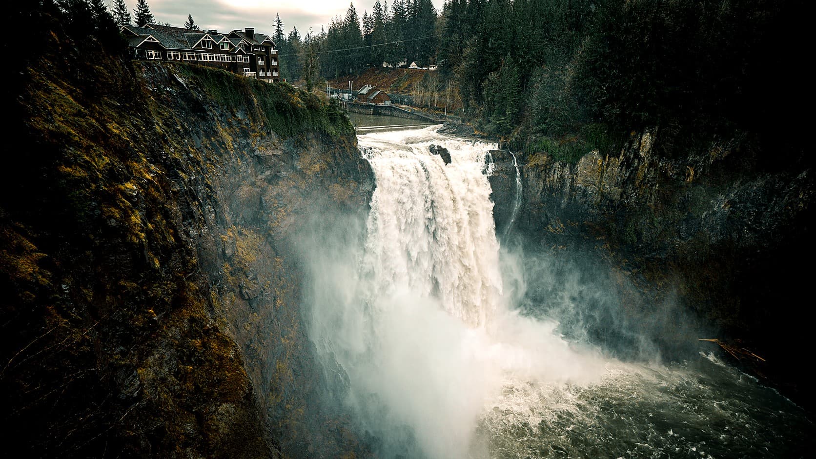 Snoqualmie Falls pours a wide white curtain into a misty gorge between dark, mossy cliffs, with the Salish Lodge on the clifftop at upper left and a low dam across the river just above the brink.