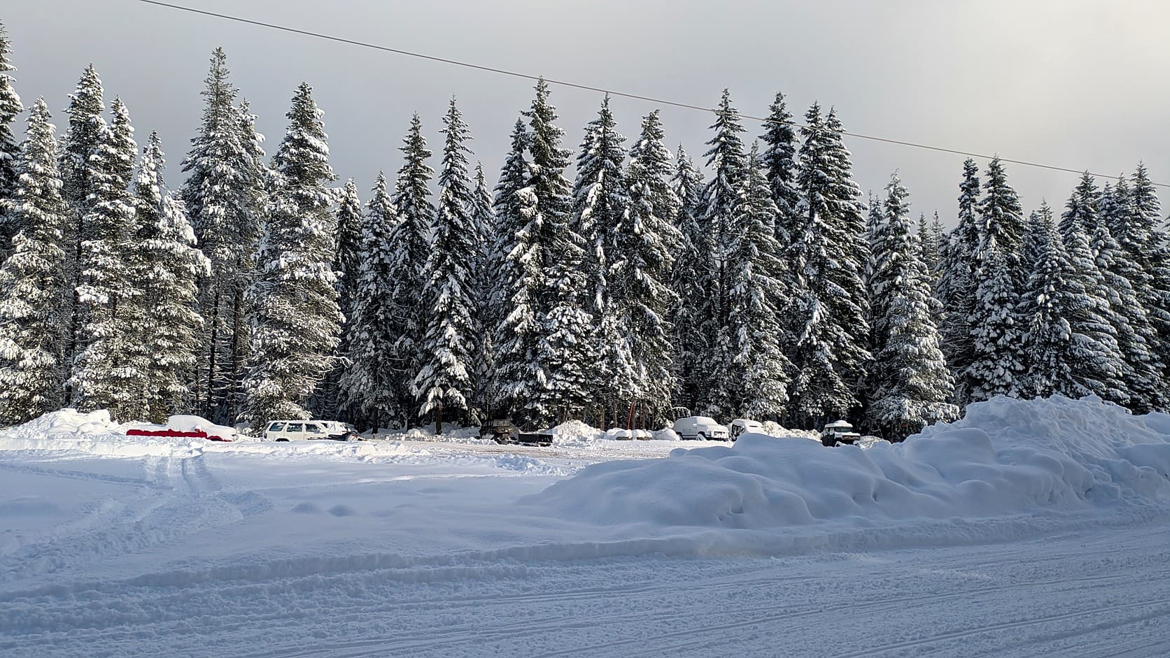Cars parked in a plowed Sno-Park lot between deep snow berms and snow-covered fir trees at Crystal Springs Sno-Park, under a gray sky crossed by a power line.