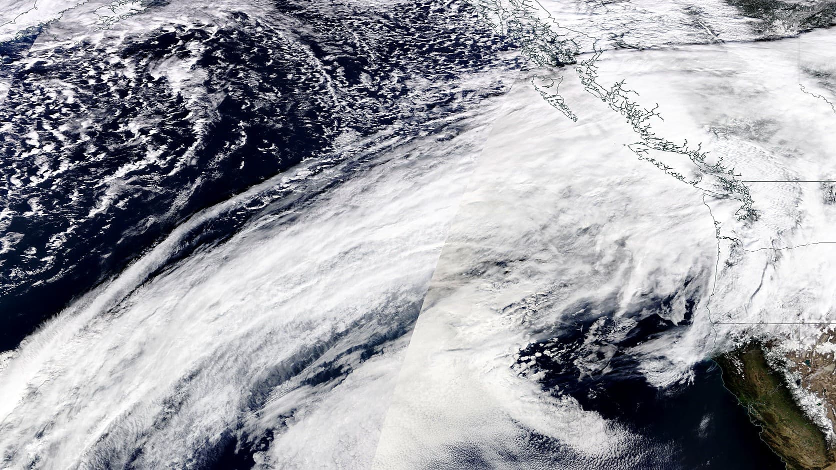A satellite image of a long white band of cloud stretching diagonally across the dark Pacific Ocean toward the coastline of British Columbia, Washington and Oregon, outlined in thin lines at the right of the frame.
