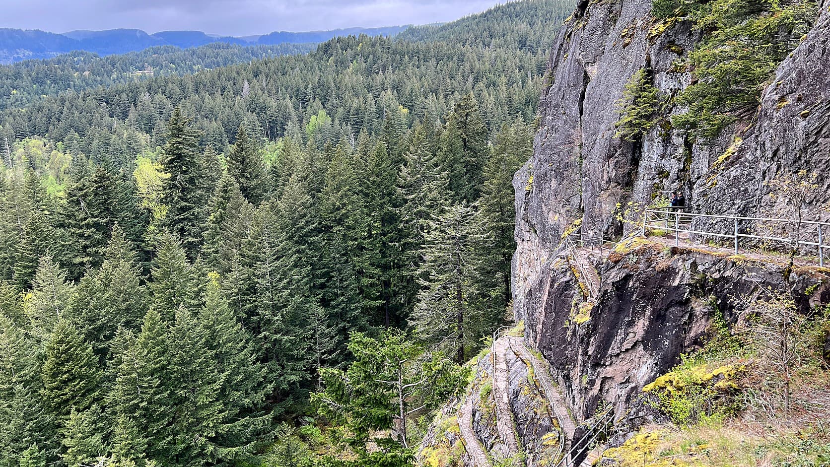 Basalt cliff face of Beacon Rock above conifer forest, with a railed trail catwalk cut into the rock on the right and forested hills in the distance under a cloudy sky.