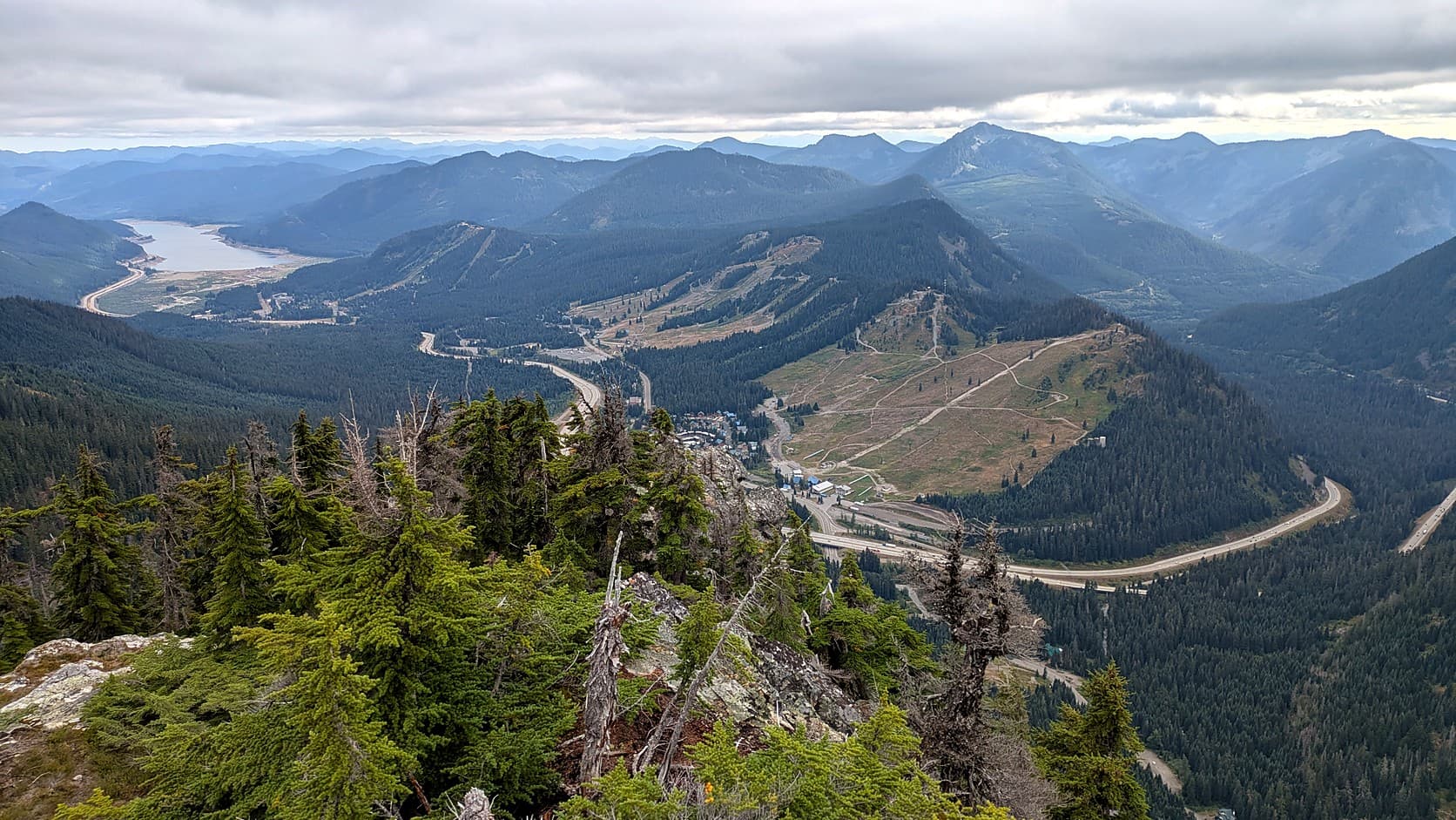 A late-summer view down onto Snoqualmie Pass from Guye Peak: bare, grass-colored ski runs on the slopes of The Summit at Snoqualmie, Interstate 90 curving through the valley below, a lake at upper left and forested ridges under an overcast sky.