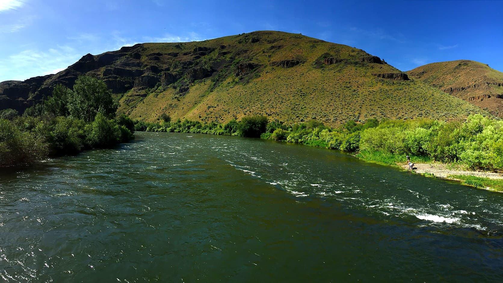 The Yakima River flowing past a green hillside with basalt outcrops under a blue sky, in the Yakima River Canyon.